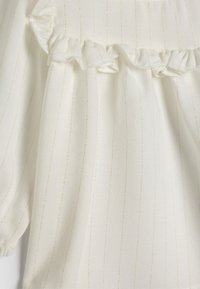 Vit blus med vertikala guldstrimmor, volangenacke och mjukt, strukturerat tyg. Löst sittande ärmar med elastiska muddar.