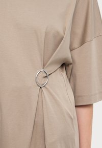 T-shirt beige oversize con maniche corte, caratterizzato da un dettaglio a nodo fissato da un anello in argento. Texture liscia, design casual.