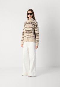 Pull en tricot beige avec un design à motif marron ; col montant et ourlet côtelé. Associé à un pantalon évasé blanc et des lunettes de soleil noires.