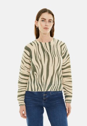 Mujer vistiendo un suéter beige y verde con rayas de cebra y jeans azules, de pie frente a un fondo blanco liso.