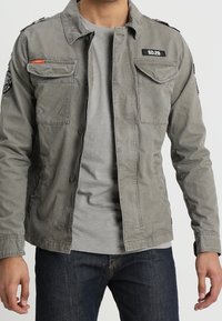 Grå bomuldsjakke med to brystlommer, knaplukning og skulderlapper. Båret over en grå skjorte og blå jeans.