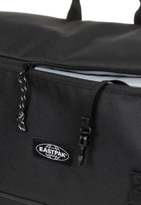 Borsa di stoffa nera con apertura con zip, superficie texturizzata, con una toppa con logo e un cordino con un motivo in bianco e nero.