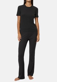 Woman standing barefoot wearing a plain black short-sleeve shirt and black pants against a white background.