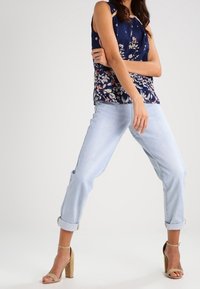 Blommig marinblå ärmlös topp tillsammans med ljusblå uppvikta jeans och beige blockklack-sandaler, vilket visar en avslappnad och vardaglig outfit.