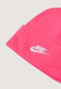 Spilgti rozā adīta vāciņš ar salocītu malu, uz kura uzgleznots balts izšūts Nike logo uz vienkārša gaiša fona.