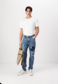 Faded blue denim jeans with patchwork detailing, paired with a plain white t-shirt. Model holding a beige bag with a crossbody strap.