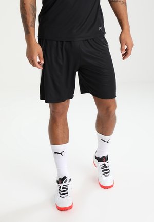Homme portant un short de sport noir, des chaussettes blanches Puma et des crampons blancs avec des semelles rouges, debout devant un fond uni.