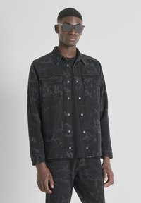 Zwarte denimjack met marmerpatroon, twee borstzakken, knoopsluiting en verstelbare manchetten. Dragen over een zwart T-shirt.
