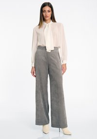 Beige blouse met een strikcollar, gecombineerd met hoog-gesneden grijze corduroy brede broek. Model draagt lichtgekleurde puntige schoenen.
