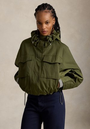 Femme aux cheveux tressés, portant une veste à capuche vert olive avec de grandes poches et un pantalon bleu marine, debout devant un fond neutre.