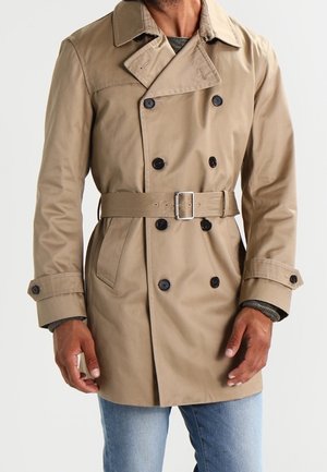 Man in een beige dubbelzijdige trenchcoat met zwarte knopen, een riem en manchetten over blauwe spijkerbroeken.