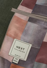 Vêtements slim fit dans des teintes de rose, pêche et lavande avec une texture douce. Présente une étiquette indiquant "NEXT TAILORING" et des instructions d'entretien.