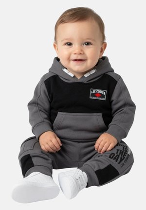 Bébé souriant assis portant une tenue à capuche grise et noire avec des chaussures blanches, le texte sur le pantalon indique "Today is the day."