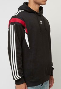 Čierna mikina s kapucňou a predným vreckom, s bielymi a červenými pruhmi na rukávoch a malým bielym logom na hrudi. Materiál z bavlneného zmesi.