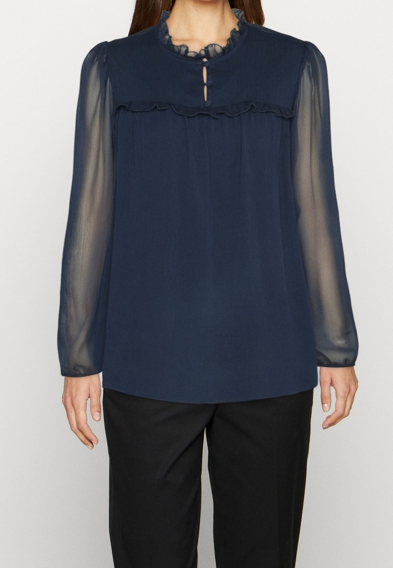 Blouse bleu marine avec manches longues transparentes, col à volants, boutons devant, portée avec un pantalon noir sur une personne debout devant un fond uni.