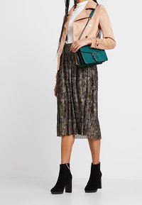 Perzikkleurige faux suede jack, witte top, geplooide midi rok met slangprint, teal handtas met ronde hardware, en zwarte suede enkellaarsjes met hak.