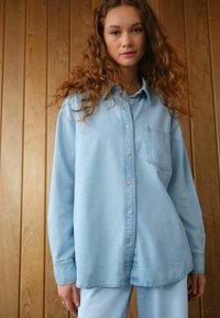 Lichtblauwe denimshirt met een knoopsluiting aan de voorkant, één borstzak en lange mouwen. Soepele textuur, relaxte pasvorm en afgeronde zoom.