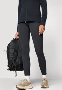 Schwarze Zip-Jacke, graue Leggings, beige Sneakers und ein schwarzer Rucksack. Strukturiertstoff auf der Jacke, elegantes Design bei Leggings und Schuhen.