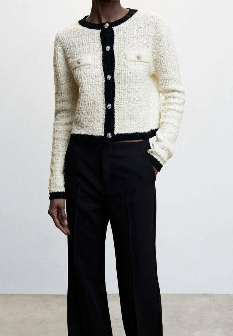 Femme portant un cardigan blanc texturé avec des bordures noires et des boutons en perles, assorti à un pantalon noir taille haute à jambes larges.