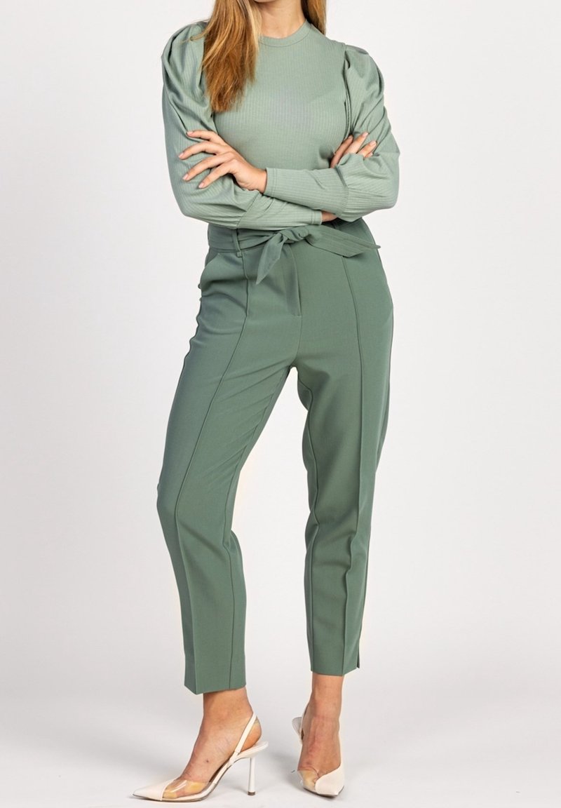 Lichtgroene geribbelde top met lange mouwen en opgepofte schouderdetails, gecombineerd met hooggetailleerde, nauwsluitende groene broek en doorzichtige schoenen met hak.