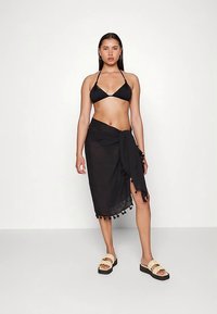 Sort bikinitop kombineret med en gennemsigtig sort slå-om-nederdel med kvastdetaljer, båret med beige slide-sandaler på en ensartet lys baggrund.