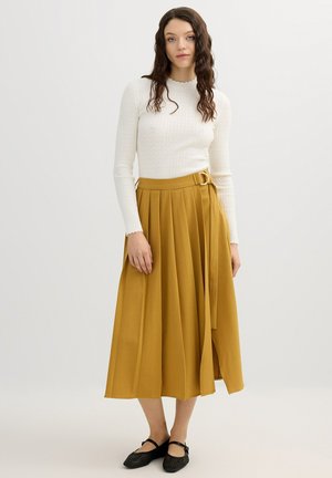 Mujer con cabello castaño ondulado, que lleva una camiseta blanca de manga larga, falda midi plisada mostaza con cinturón y zapatos planos negros, de pie sobre un fondo blanco.