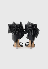 Black high-heeled sandals with large leather bows at the back and thin stiletto heels against a plain white background.