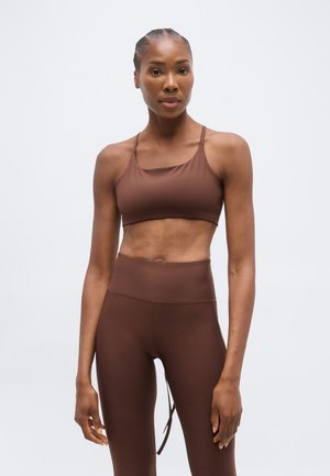 Atletická žena s pletenými vlasmi v hnedom športovom podprsenke a legínach s vysokým pásom stojaca pred jednofarebným svetlým pozadím.