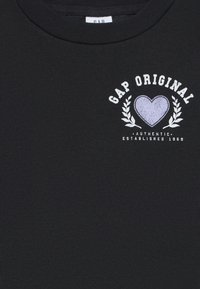Maglietta nera con il testo "GAP ORIGINAL" sopra un cuore viola con rami di alloro bianchi e "AUTHENTIC ESTABLISHED 1969" sotto.