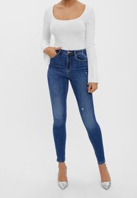 Korkeavyötäröiset siniset farkut, joissa on kulutettu yksityiskohta, yhdistettynä valkoiseen ribattuun pitkähihanteeseen ja hopeisiin korkokenkiin.