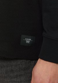 Schwarzer Sweatshirt mit gerippten Säumen und Bund, versehen mit einem aufgenähten S.Oliver-Label. Der Stoff wirkt glatt und hat ein einfaches, klares Design.