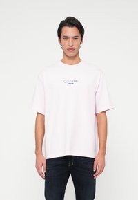 Mann mit einem hellrosa Calvin Klein Jeans T-Shirt und dunkelblauen Jeans, der vor einem schlichten weißen Hintergrund steht.