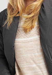 Musta toppatakki vaalean beigen neuleen päällä, jossa on teksturoitu kuvio, pehmeä neulottu kangas ja pyöreä pääntie.