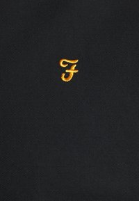 Tissu noir présentant de petites lettres minuscules stylisées "fj" brodées en fil jaune doré au centre.