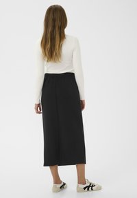 Femme aux cheveux longs vêtue d'un haut blanc à manches longues, d'une jupe midi noire et de baskets blanches avec des accents noirs, se tenant de dos.