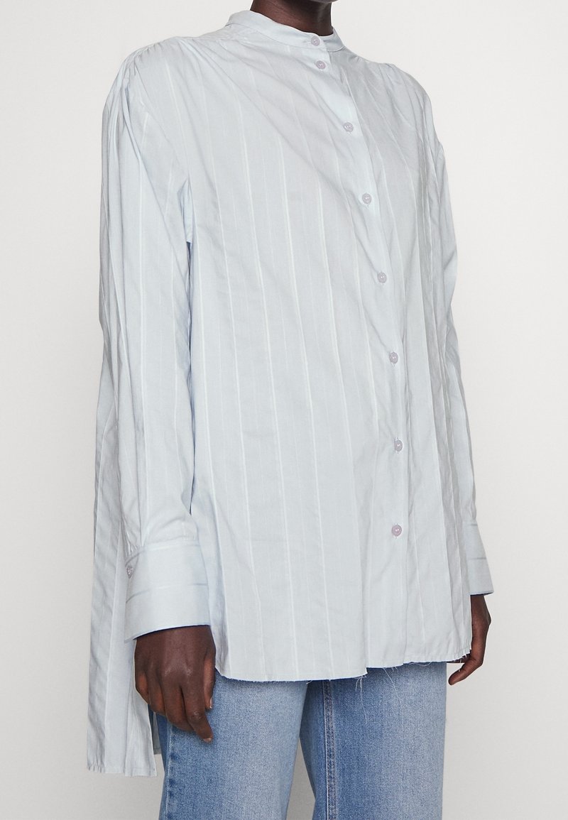 Osoba oblečená v volné, světle modré pruhované košili s mandarin límcem a světle modrých džínách na jednobarevném pozadí.