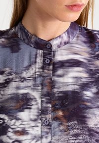 Kragbluse aus Seide mit einem verschwommenen abstrakten Muster in Lila-, Grau- und Brauntönen. Verfügt über eine Knopfverschluss und eine weiche Textur.