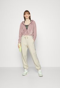 Sudadera con capucha cortada en color malva, pantalones jogger beige con cintura ajustable, combinados con zapatillas verdes claras y sosteniendo una botella de agua color lima.