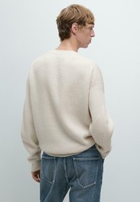 Giovane in un maglione a coste crema e jeans blu, in una posa rilassata, di spalle contro uno sfondo chiaro e uniforme.