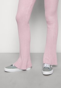 Calças flared de malha canelada rosa, combinadas com ténis de lona cinzentos. As calças apresentam uma textura elástica, enquanto os ténis têm atacadores brancos.