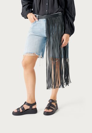 Persona que lleva shorts de mezclilla claros y desgarrados, blusa negra abotonada, cinturón negro con flecos y sandalias negras abiertas, de pie frente a un fondo liso.