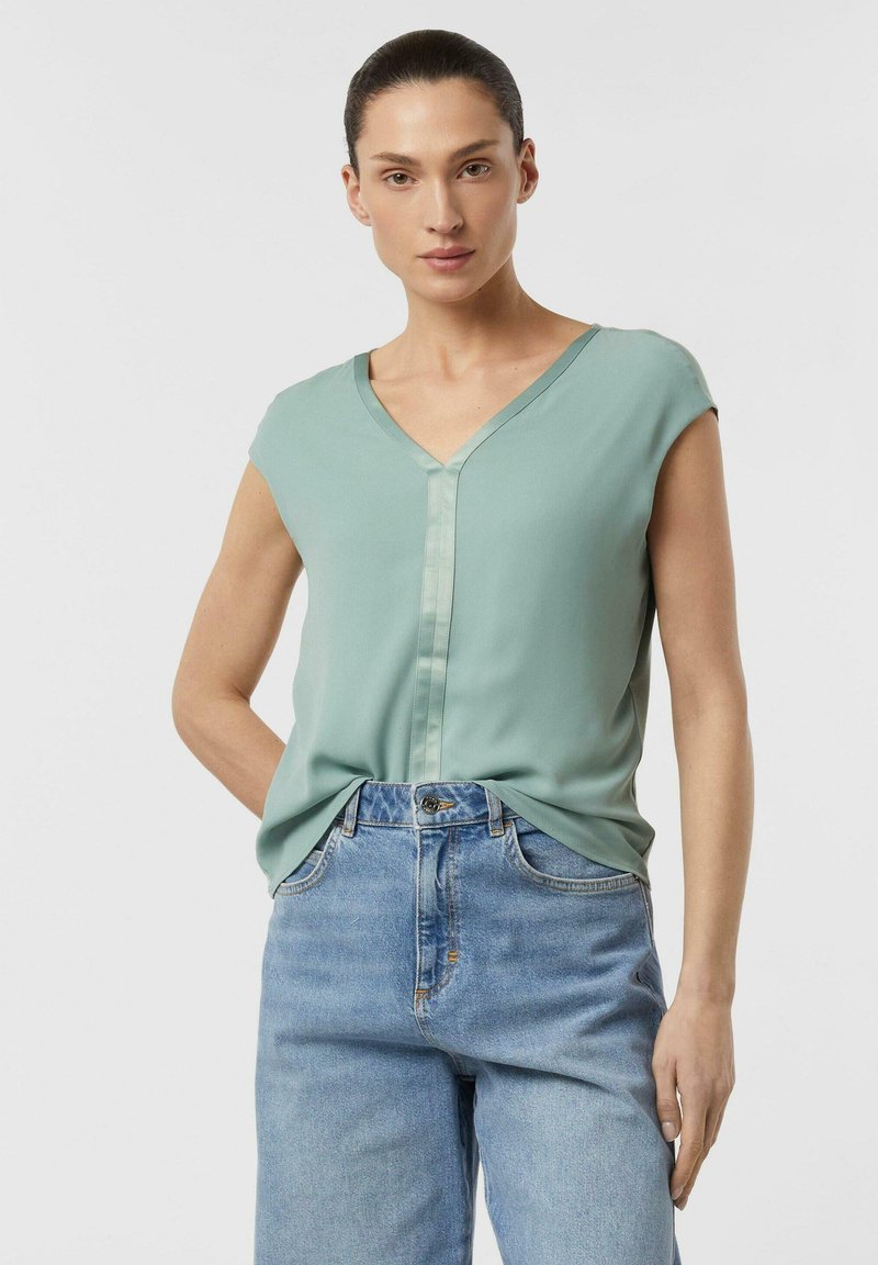 Femme aux cheveux foncés attachés portant un chemisier vert clair à col en V rentré dans un jean taille haute bleu clair, sur fond blanc.