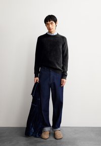Schwarzer Rundhalsausschnitt-Pullover, marineblaue weit geschnittene Jeans, braune Slipper, hält eine marineblaue Jacke. Minimalistisches Design, weiche Texturen, lässiger Look.