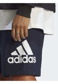 Námornícke šortky adidas s veľkým bielym logom, bavlnený materiál, elastický pás, kombinované s bielym vrstveným topom. Ruka s prsteňom viditeľná.
