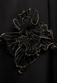 Black fabric with a raised, black floral embellishment outlined in metallic gold stitching, featuring intricate layered petals and fine detailing.