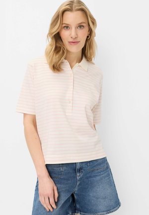 Blonde vrouw draagt een wit polo shirt met korte mouwen met lichtroze strepen en blauwe spijkershorts, staand tegen een witte achtergrond.