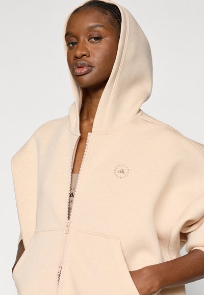 Μπεζ υπερμεγέθη φούτερ με φερμουάρ, μεγάλη κουκούλα, μπροστινή τσέπη και λογότυπο στο στήθος. Μαλακή υφή και άνετη εφαρμογή.