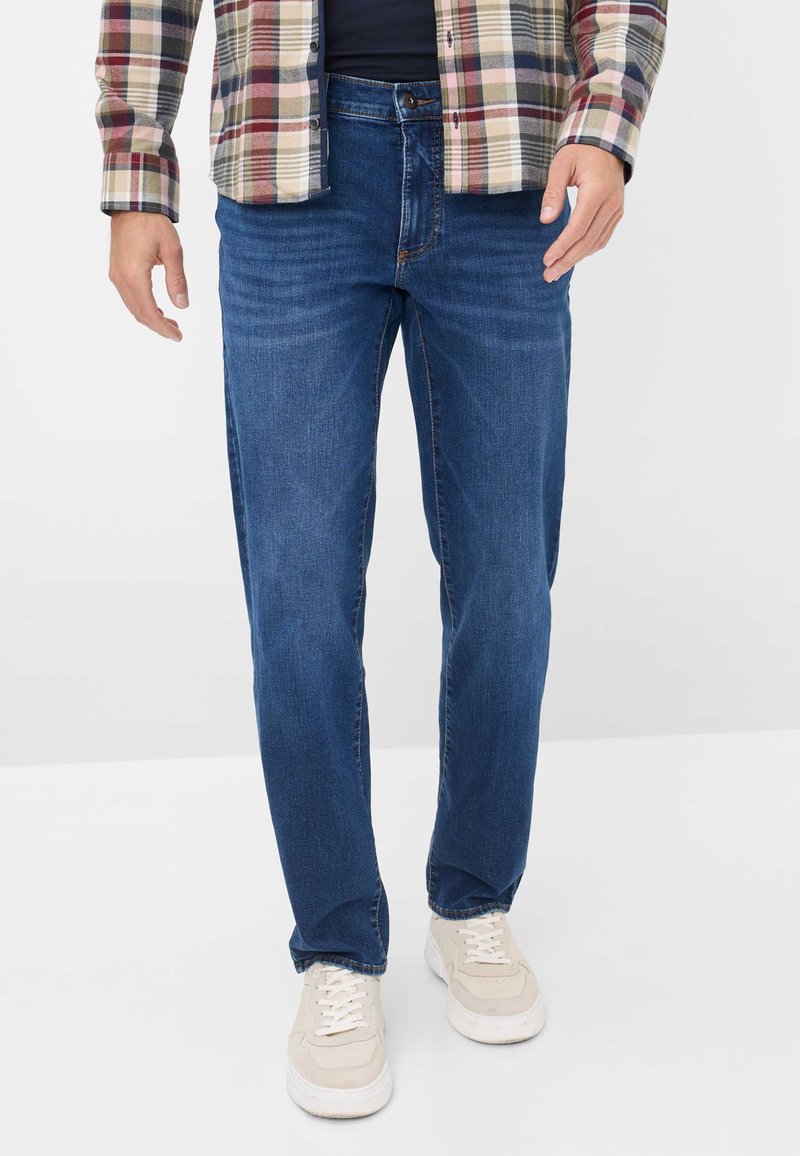 Jean droit bleu foncé avec fermeture à bouton et design à cinq poches, porté avec une chemise à carreaux et des baskets beiges.