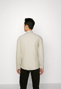 Camicia a maniche lunghe color beige chiaro con sottili righe verticali, colletto, polsini abbottonati e vestibilità rilassata. Indossata sopra pantaloni scuri.