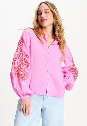 Vrouw draagt een roze blouse met knopen en rode bloemrijke borduursels op opgepofte mouwen en lichtblauwe jeans, staand met één hand in de zak.